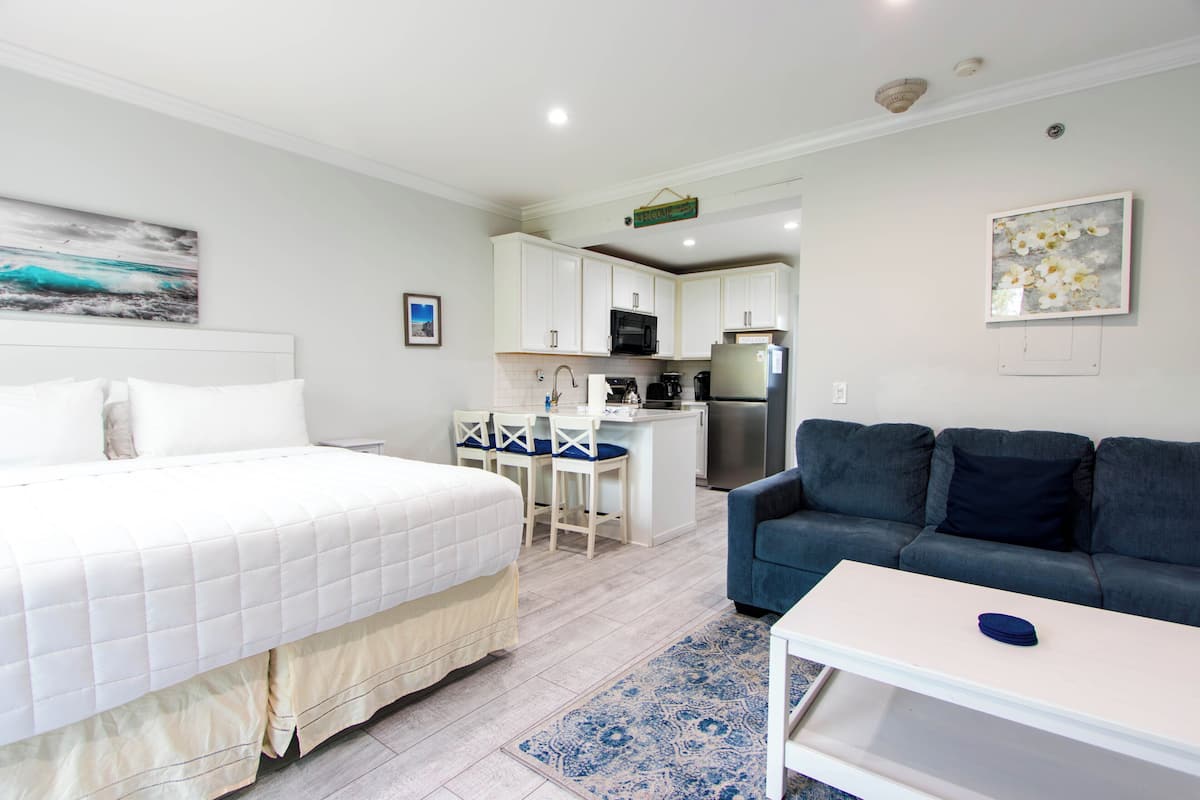 A bedroom with a bed, a blue sofa, a small table, and a kitchen area in the background.