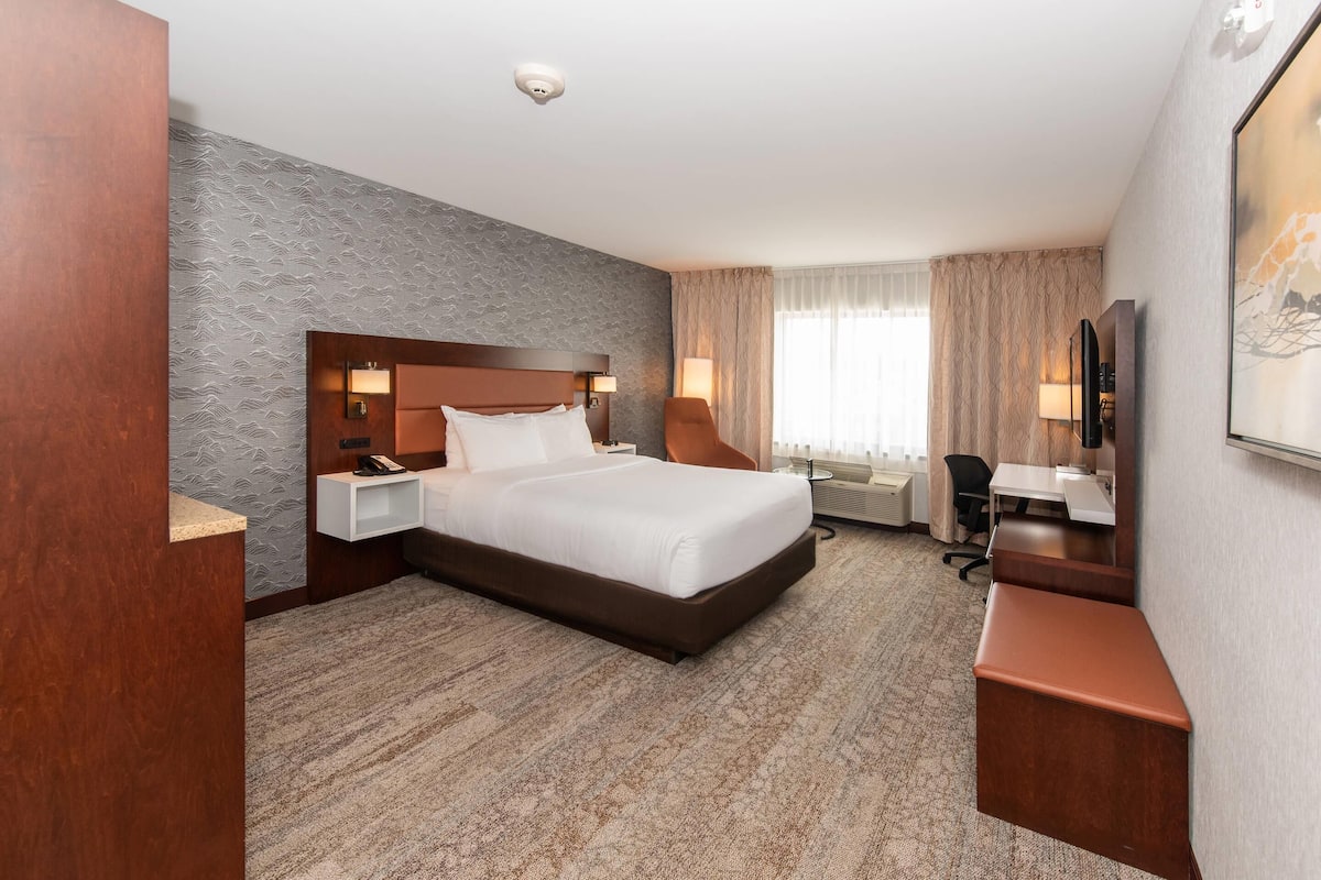 Quarto de hotel com uma cama grande, uma escrivaninha, uma cadeira, televisão e uma janela com cortinas.