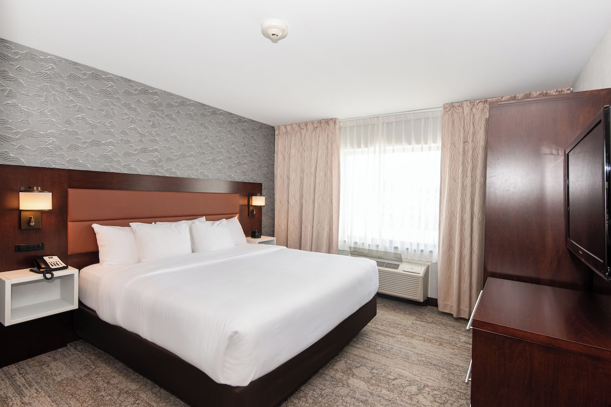 Quarto de hotel com uma cama grande, cabeceira de madeira, mesa de cabeceira com telefone, televisão e janela com cortinas.