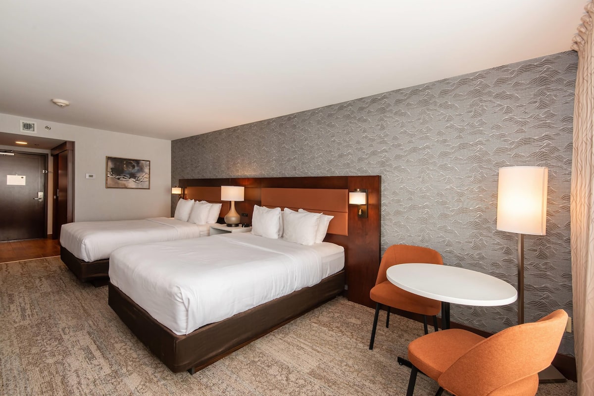 Quarto de hotel com duas camas, uma mesa pequena, uma cadeira e parede estampada.