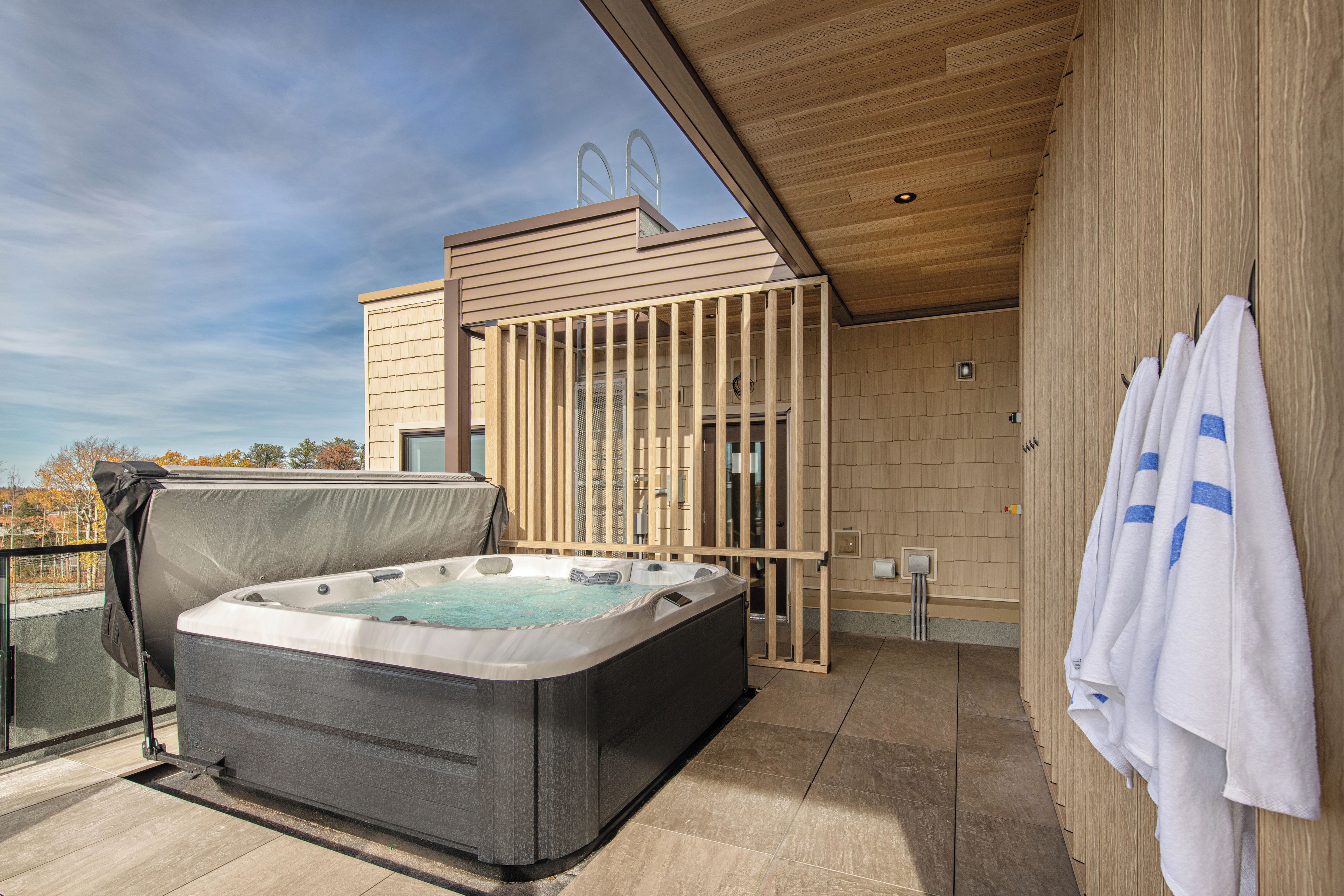 A modern outdoor hot tub area with a wooden deck, a covered seating area, and a view of the surrounding landscape.