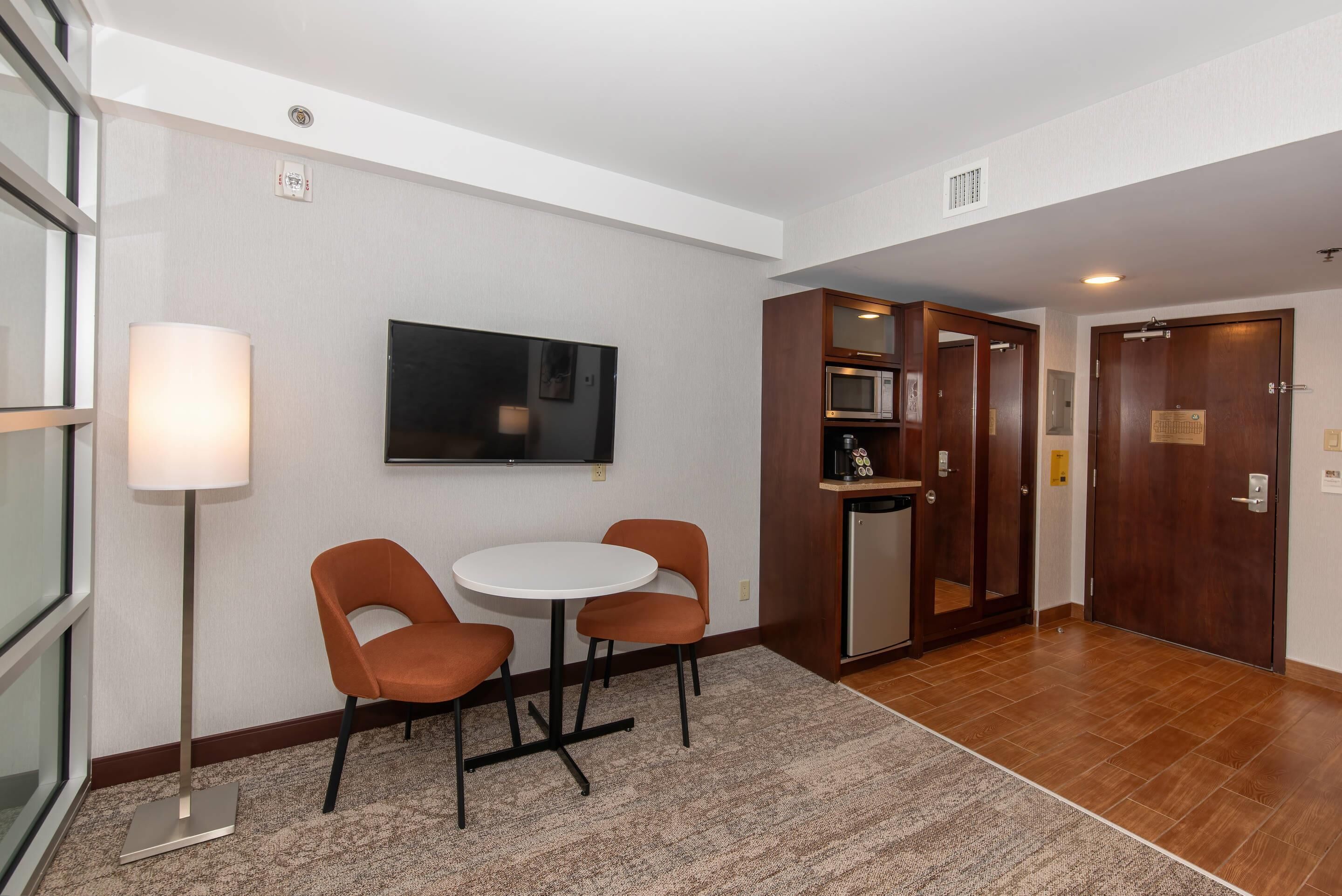 A hotel lobby with a flat-screen TV, a round table with two chairs, a microwave, and a wooden cabinet.