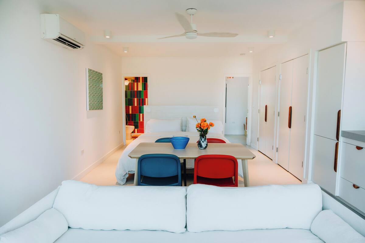 A modern hotel room with a white sofa, a dining table with blue and red chairs, a ceiling fan, and a colorful artwork on the wall.
