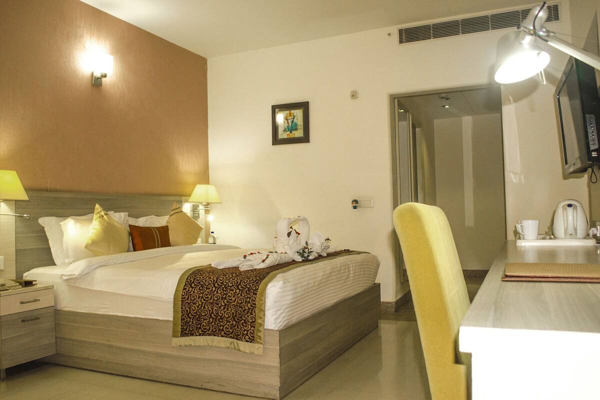 A hotel room with a bed, bedside lamps, a chair, a desk with a kettle, and a television.