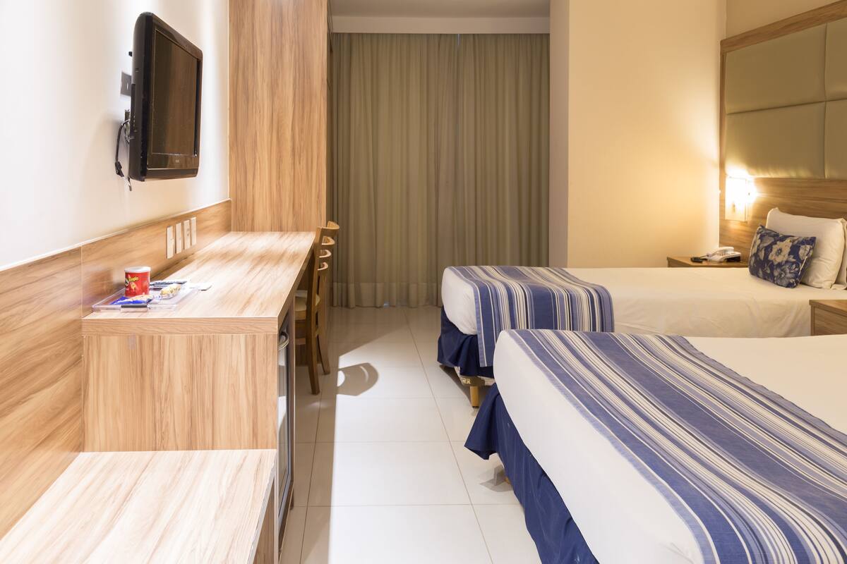 A hotel room with two beds, a desk, a TV, and a window with curtains.
