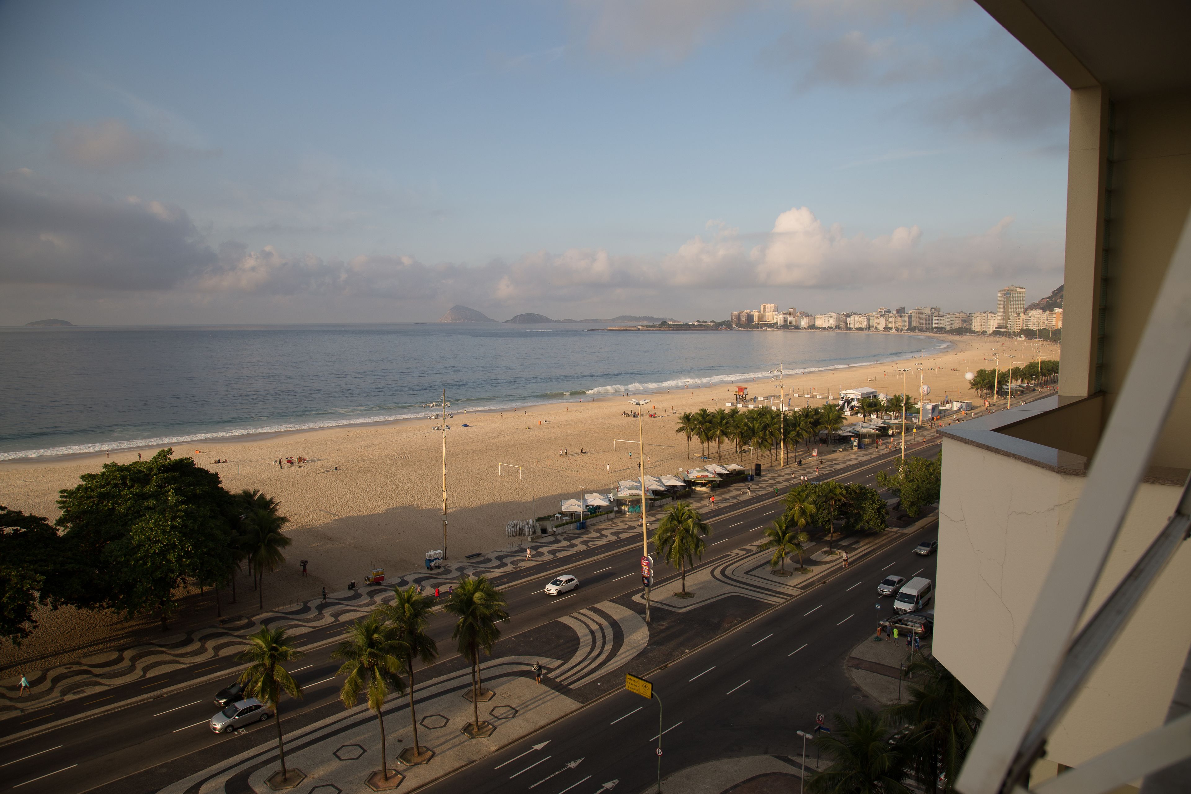 Vista de la playa desde un lugar elevado, que incluye una carretera con palmeras y vehículos, y un horizonte lejano con edificios.
