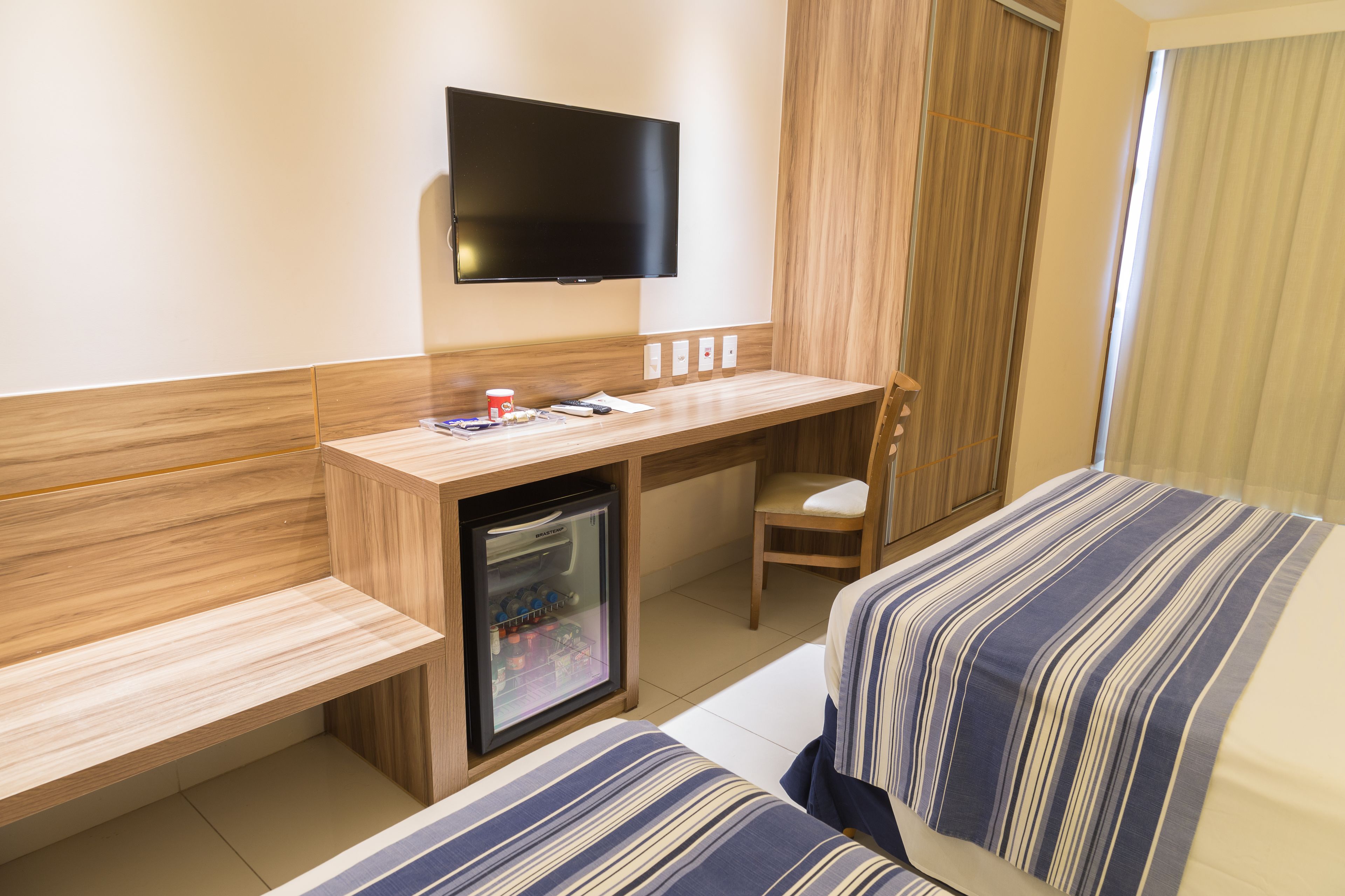Habitación de hotel con un escritorio de madera, televisor montado en la pared, un mini refrigerador, una silla y dos camas con ropa de cama a rayas.