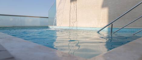 Ein Schwimmbecken mit Wasserspiel, umgeben von einer gefliesten Terrasse und einem Metallgeländer.