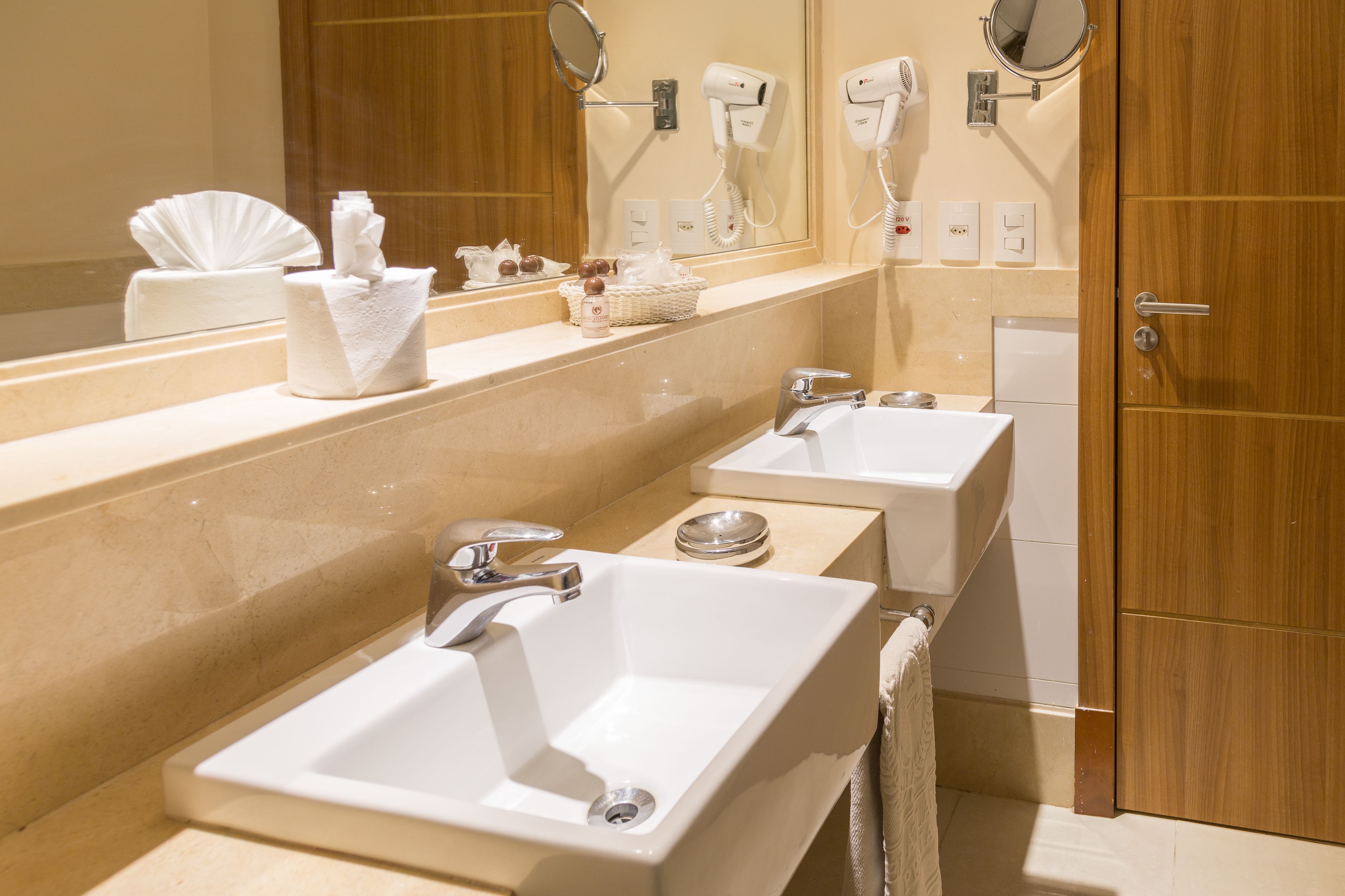 A hotel bathroom with two sinks, a mirror, a hairdryer, and a towel rack.