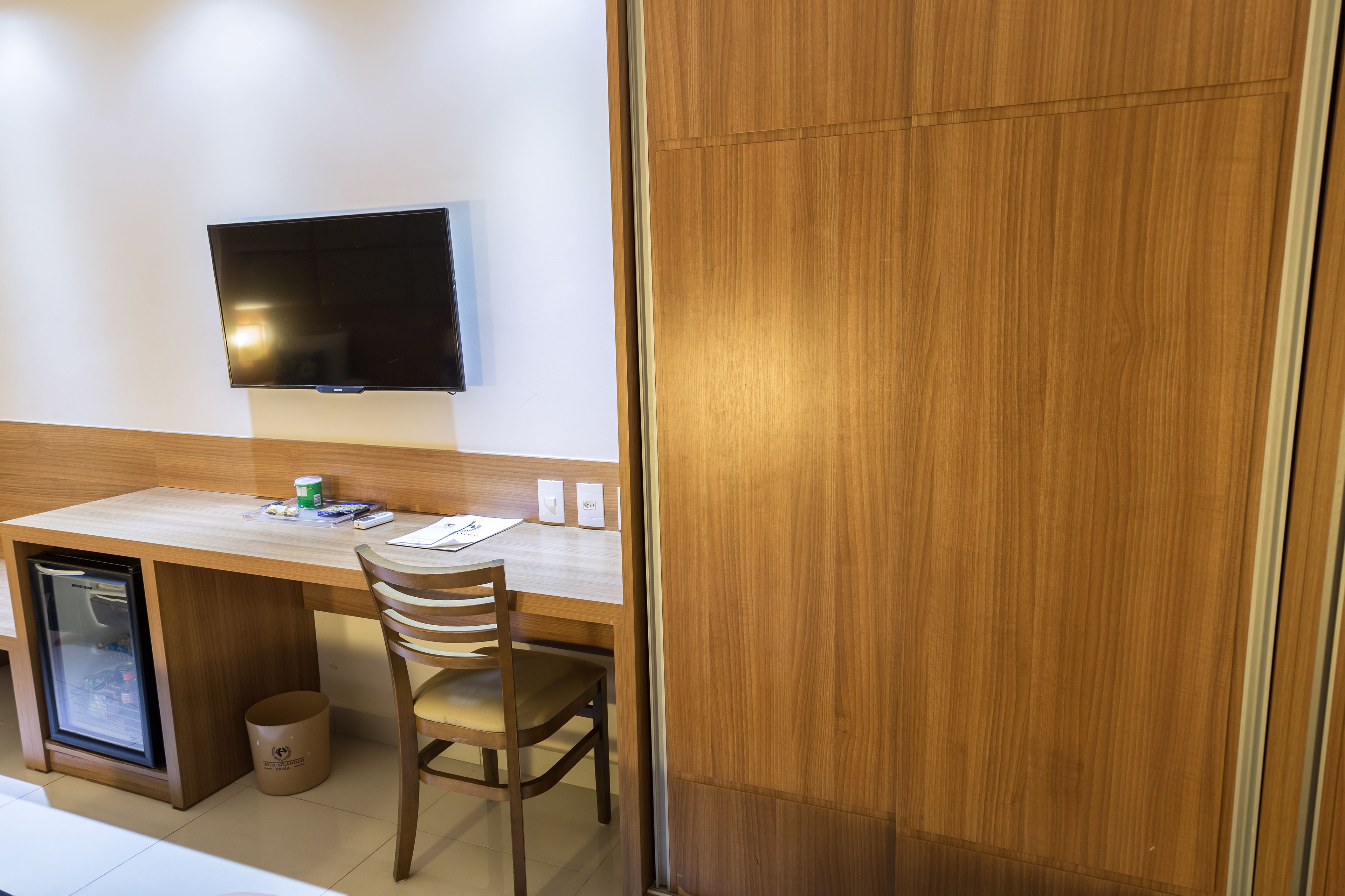 Een compacte hotelkamer met een houten bureau, een aan de muur bevestigde televisie, een kleine koelkast en een afvalbak.