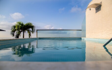 A clear blue swimming pool with a tiled edge, surrounded by a light-colored wall and a palm tree.