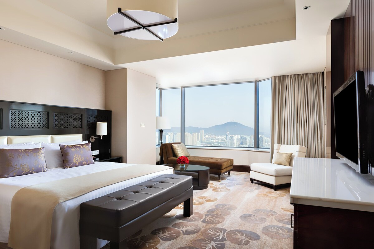 A hotel room with a large bed, a seating area with a sofa and armchair, a flat-screen TV, and a city view through large windows.