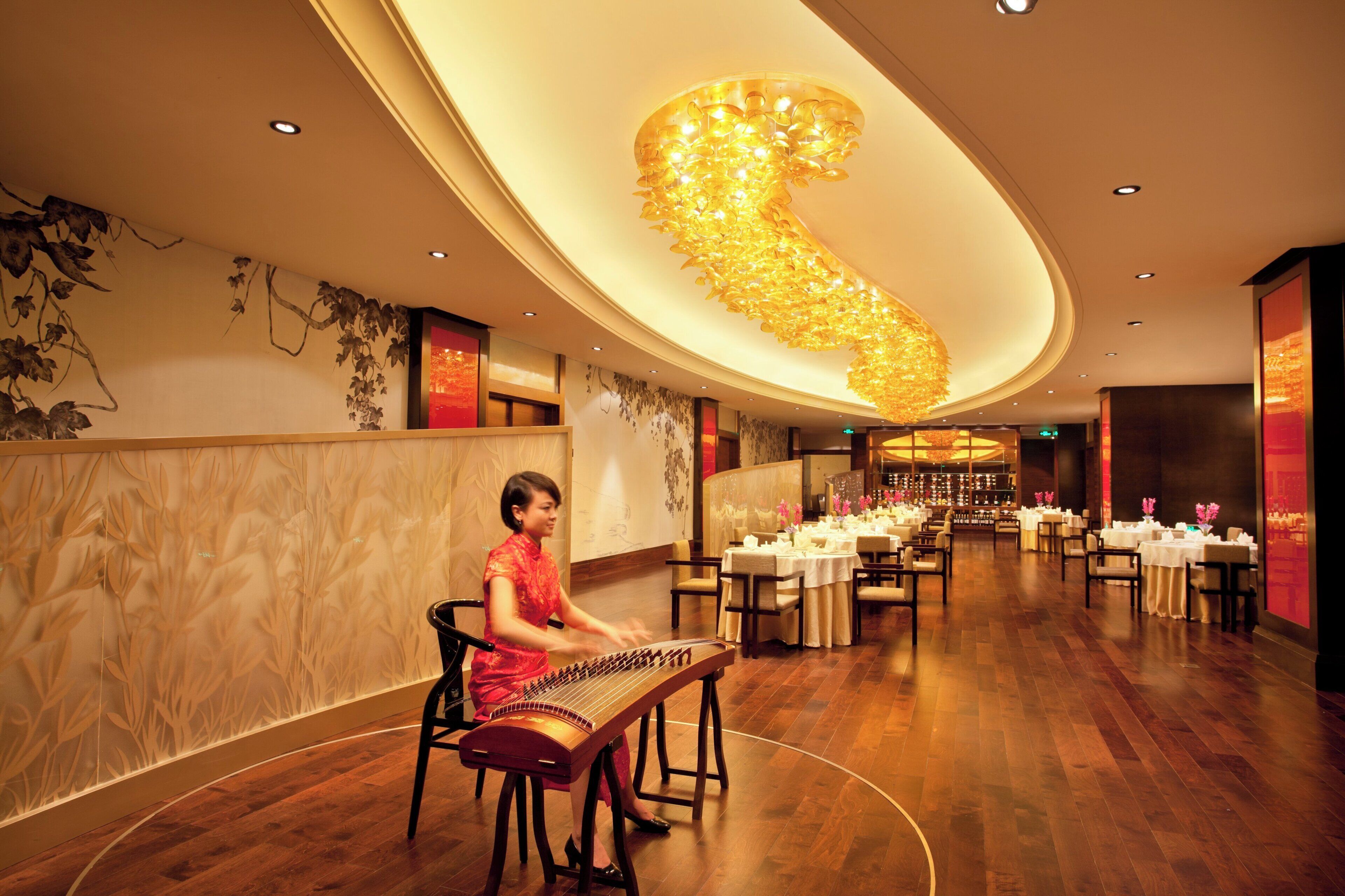 A woman playing a traditional stringed instrument in a well-lit room with a large decorative ceiling feature.