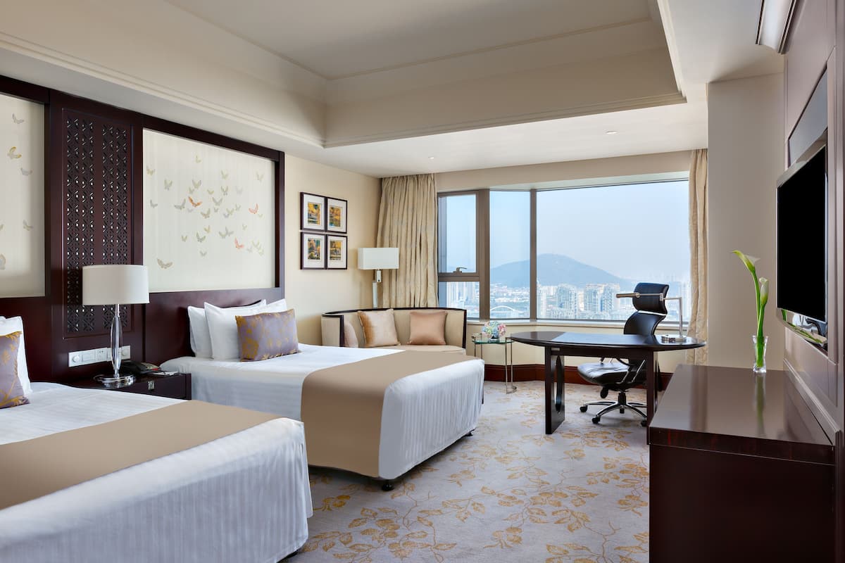 A hotel room with two beds, a desk, a chair, a TV, and a large window with a city view.