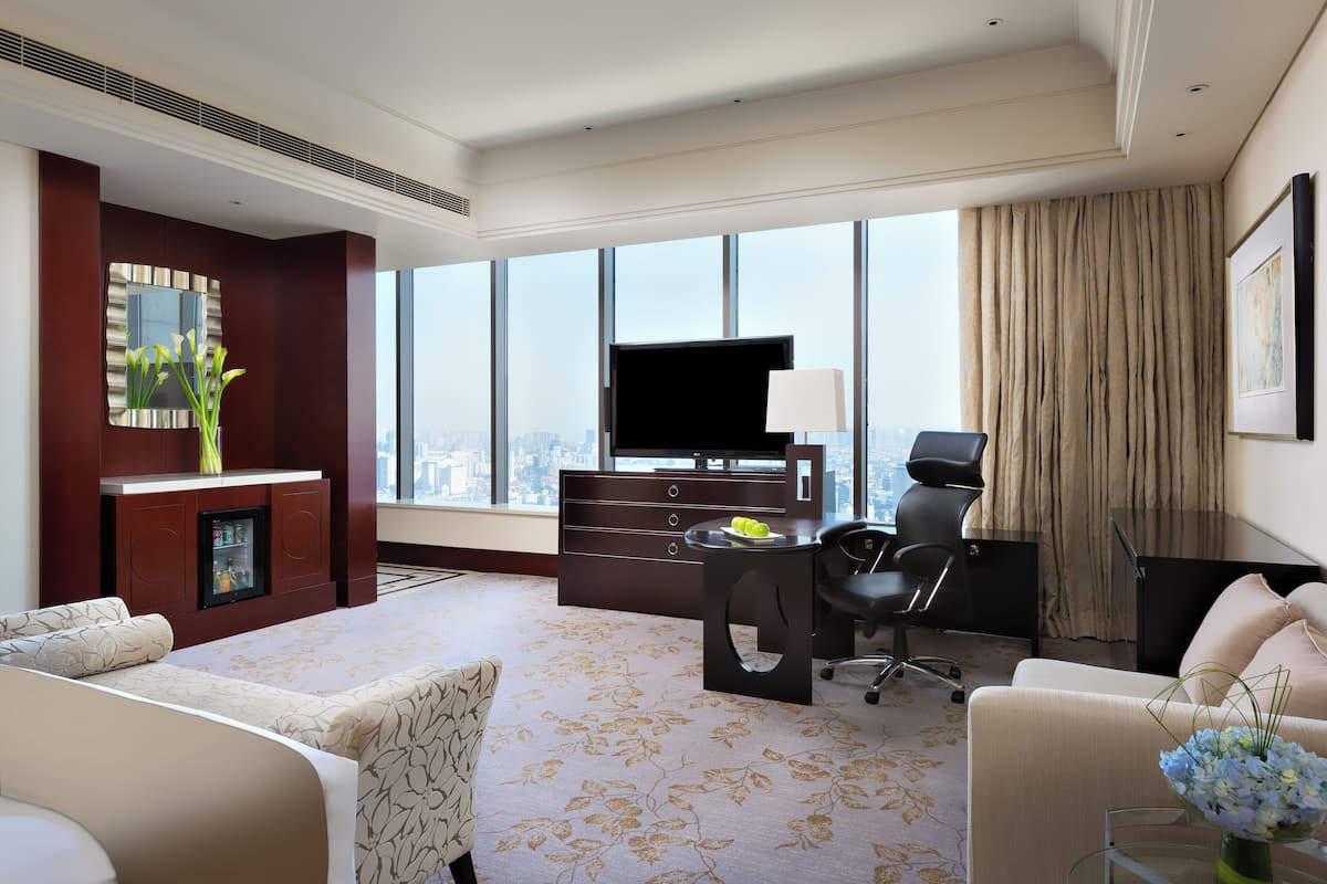 A hotel room with a large window, a flat-screen TV, a desk with a chair, and a sofa.
