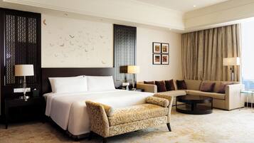 A hotel room with a large bed, a sofa, a chair, and a coffee table. There are wall decorations and a view of the city.