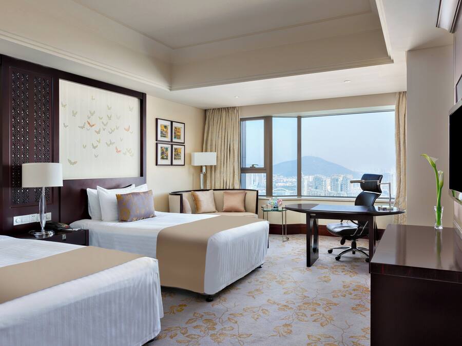 Habitación de hotel con dos camas, un escritorio, una silla, un televisor y un amplio ventanal con vista a la ciudad.