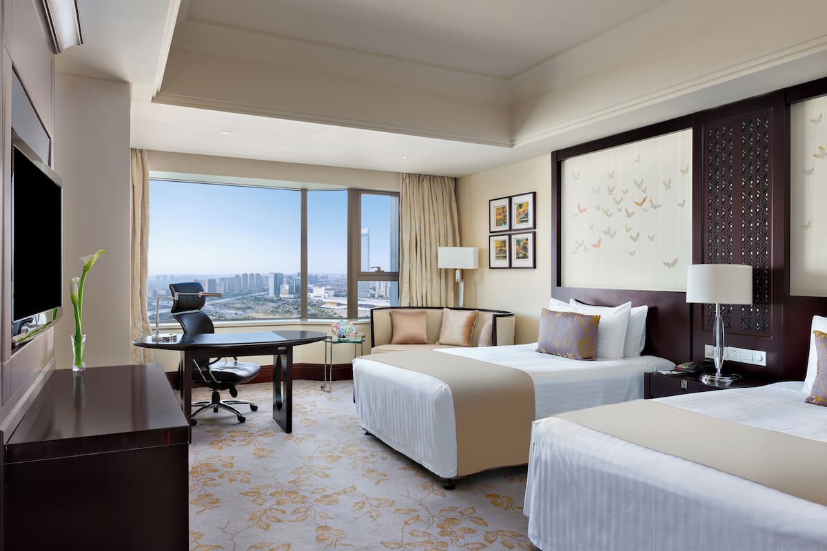 A hotel room with two beds, a desk, a TV, and a large window offering a city view.