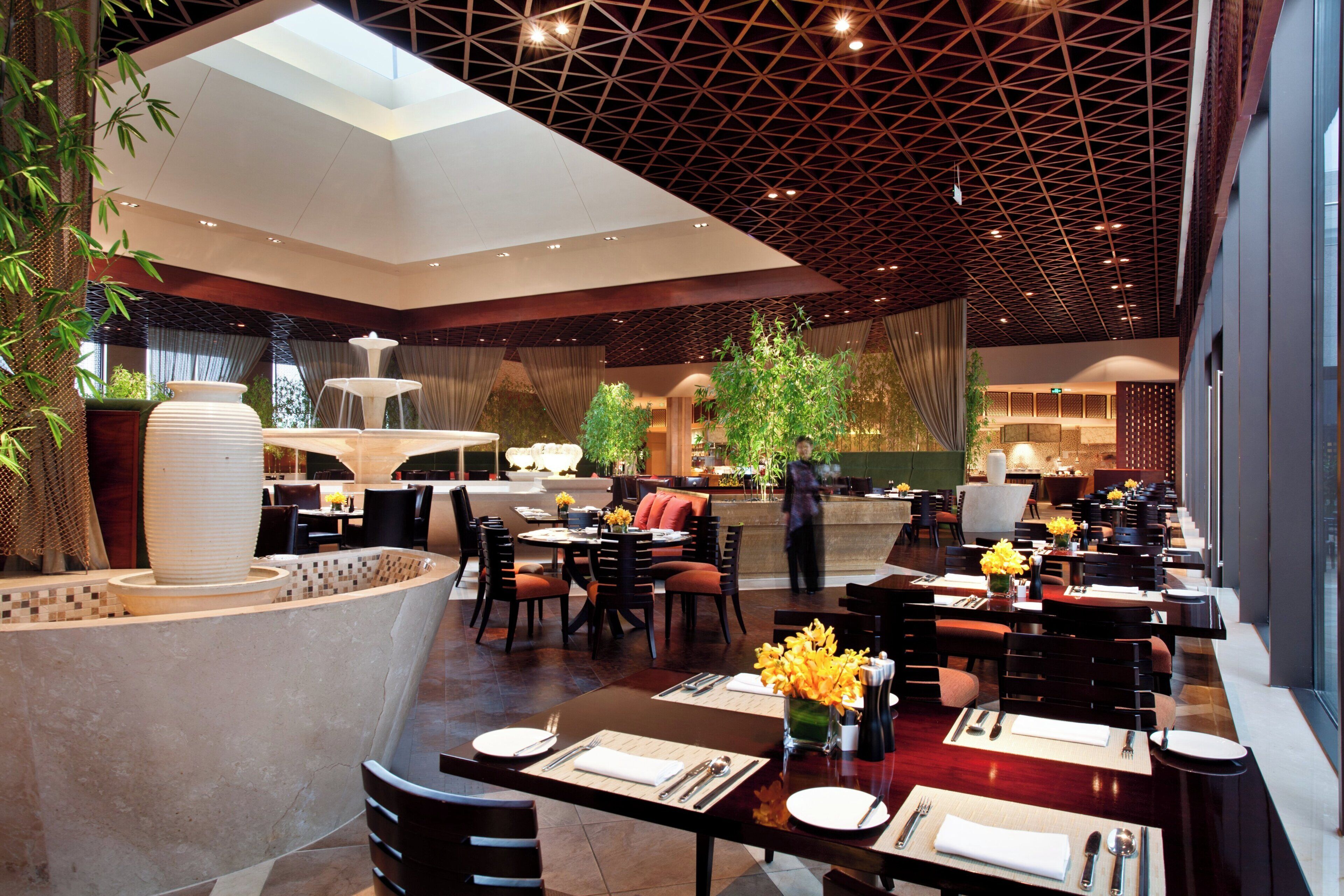 Ein modernes Restaurant mit hoher Decke, geometrischem Deckenmuster und einer zentralen Wasserspüle. Die Tische sind mit weißen Tischdecken und gelben Blumen gedeckt.