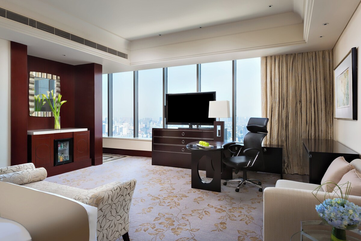 A hotel room with a large window, a flat-screen TV, a desk with a lamp, a chair, and a sofa.