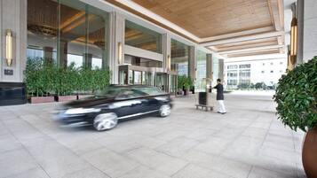 A modern building entrance with a car passing by, large glass windows, and a person standing near a kiosk.