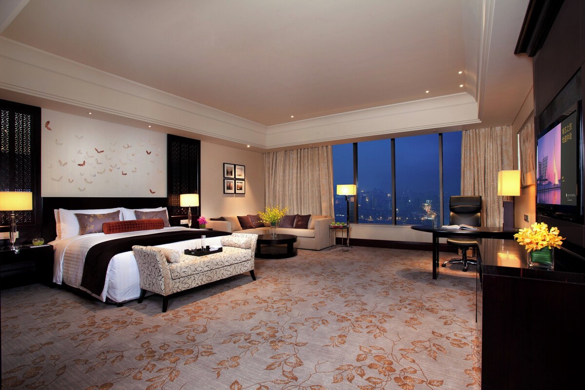 A hotel room with a large bed, a sofa, a desk, and a view of the city at night.