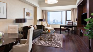 A modern hotel room with a large window offering a city view, a sofa, a dining table, and a TV.