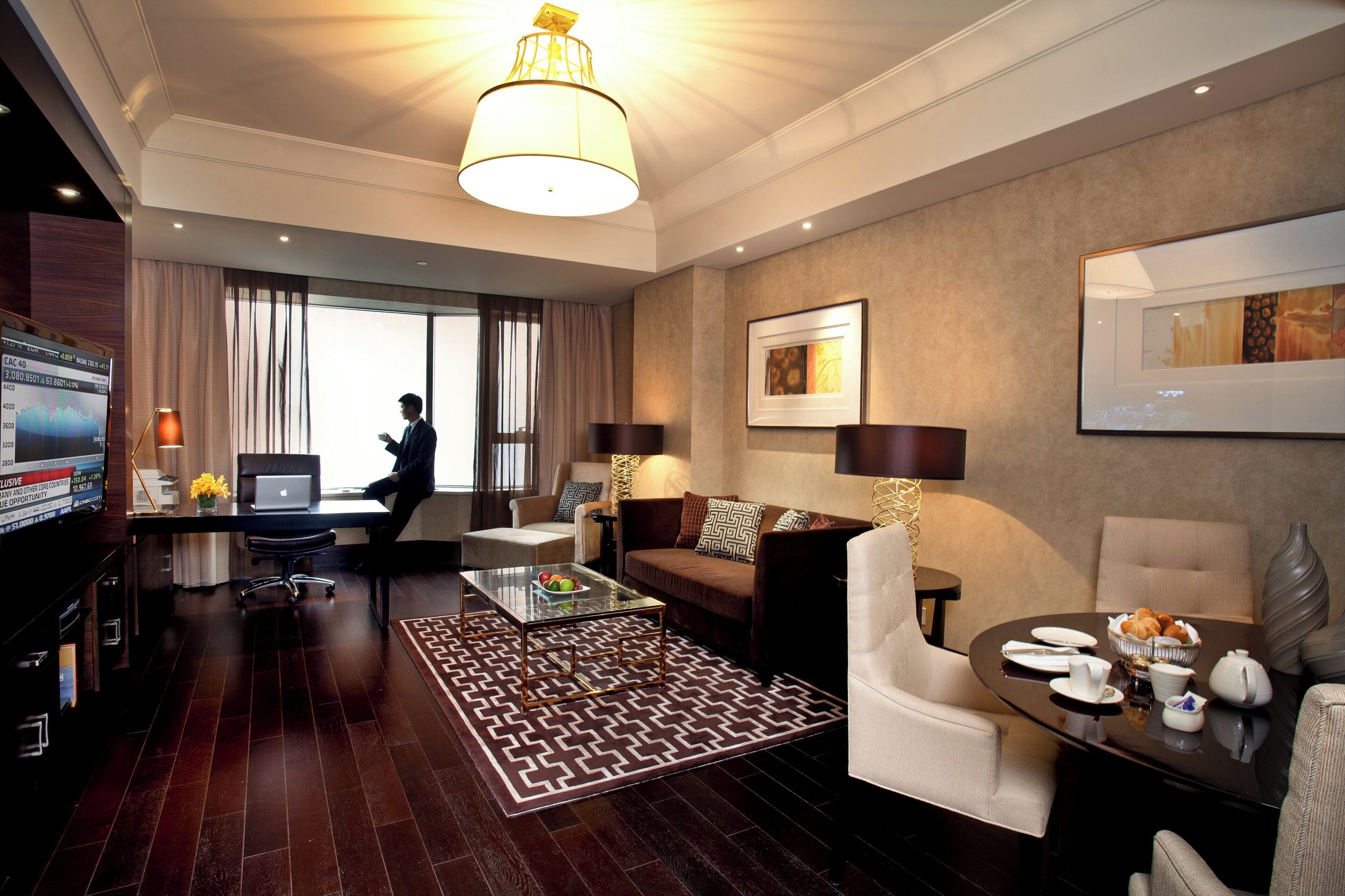 Et moderne hotelværelse med et stort vindue, et fladskærms-tv, en bærbar computer, et spisebord med en vase og en sofa.