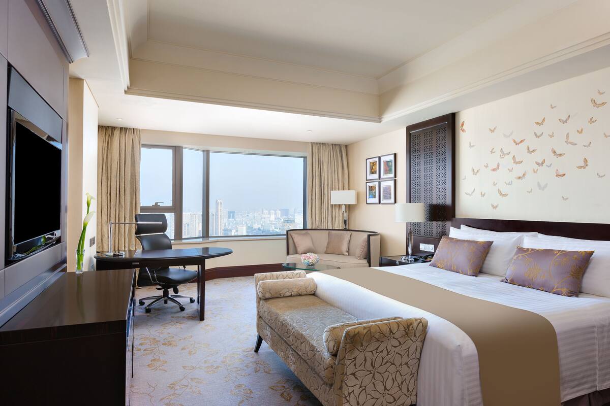 A hotel room with a large bed, a desk, a chair, a TV, and a view of the city.