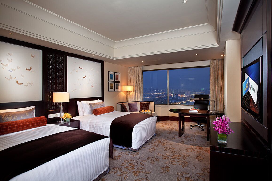Habitación de hotel con dos camas, un escritorio, televisor y una ventana grande con vista a la ciudad.
