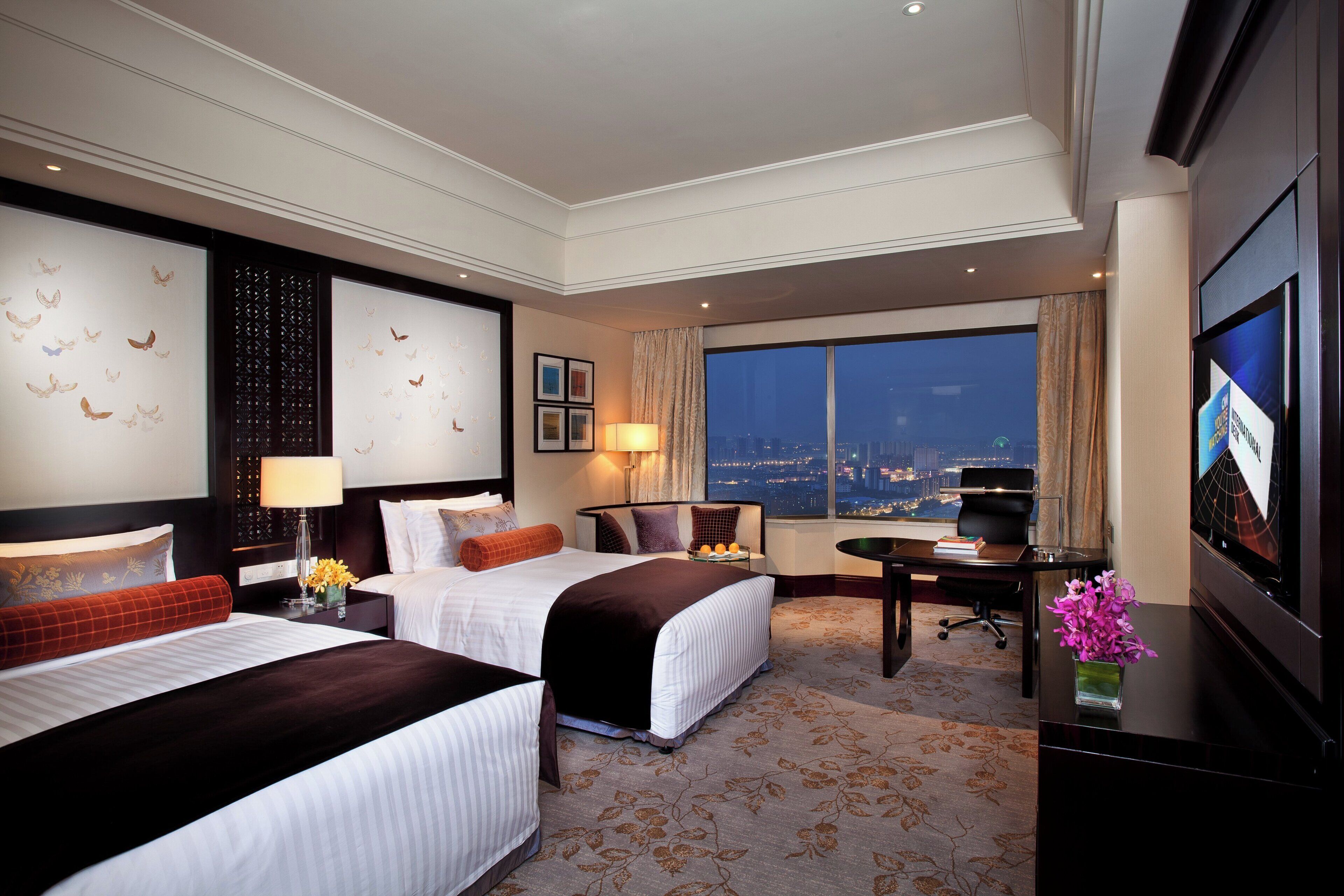 A hotel room with two beds, a desk, a television, and a large window with a city view.