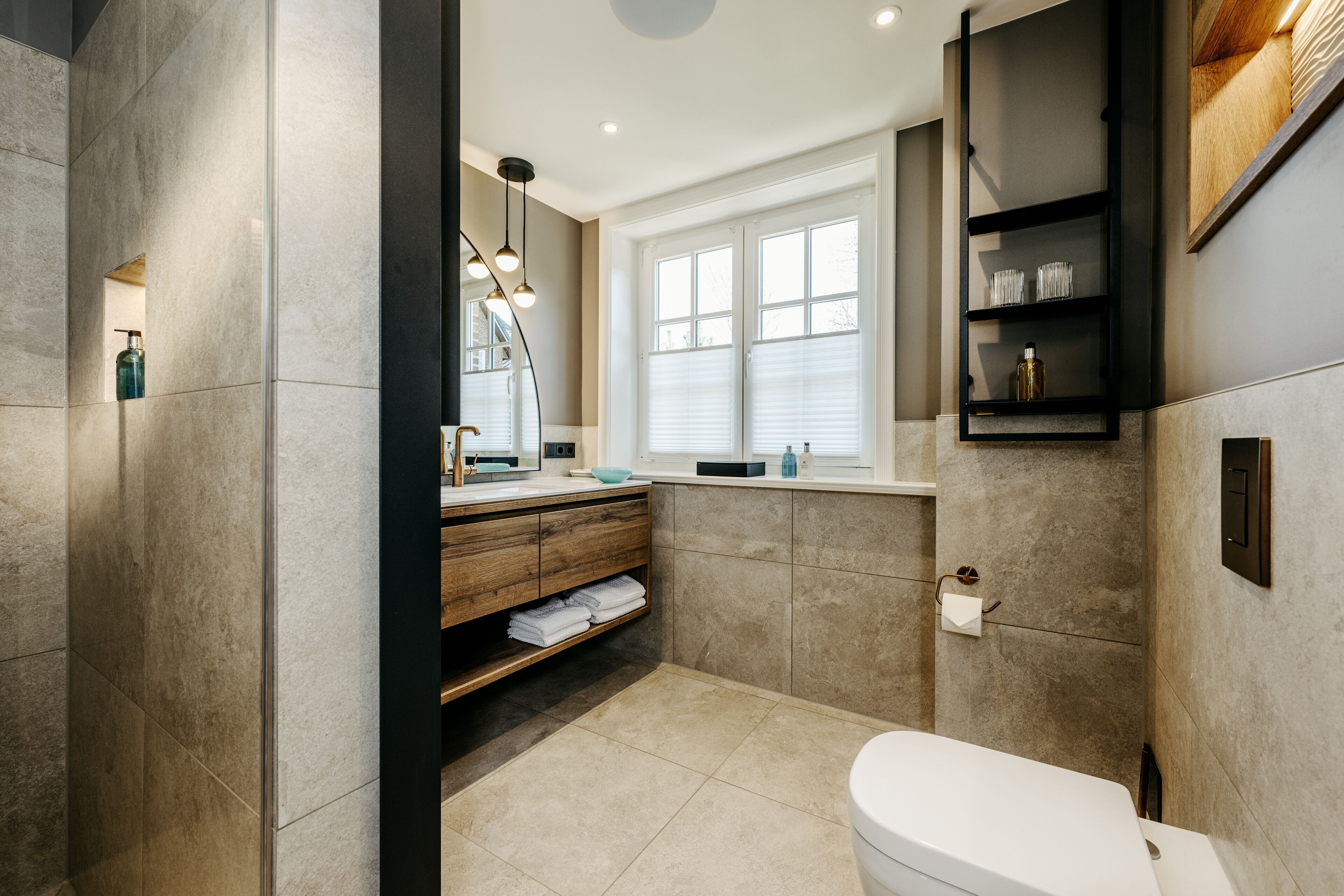 A modern bathroom with a toilet, sink, and vanity. There is a window with blinds, a shelf with toiletries, and a mirror above the sink.