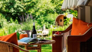 A wicker chair with a book, a bottle of wine, and a glass of wine on a table in a garden setting. - Relais & Châteaux Landhaus Stricker (Sylt)