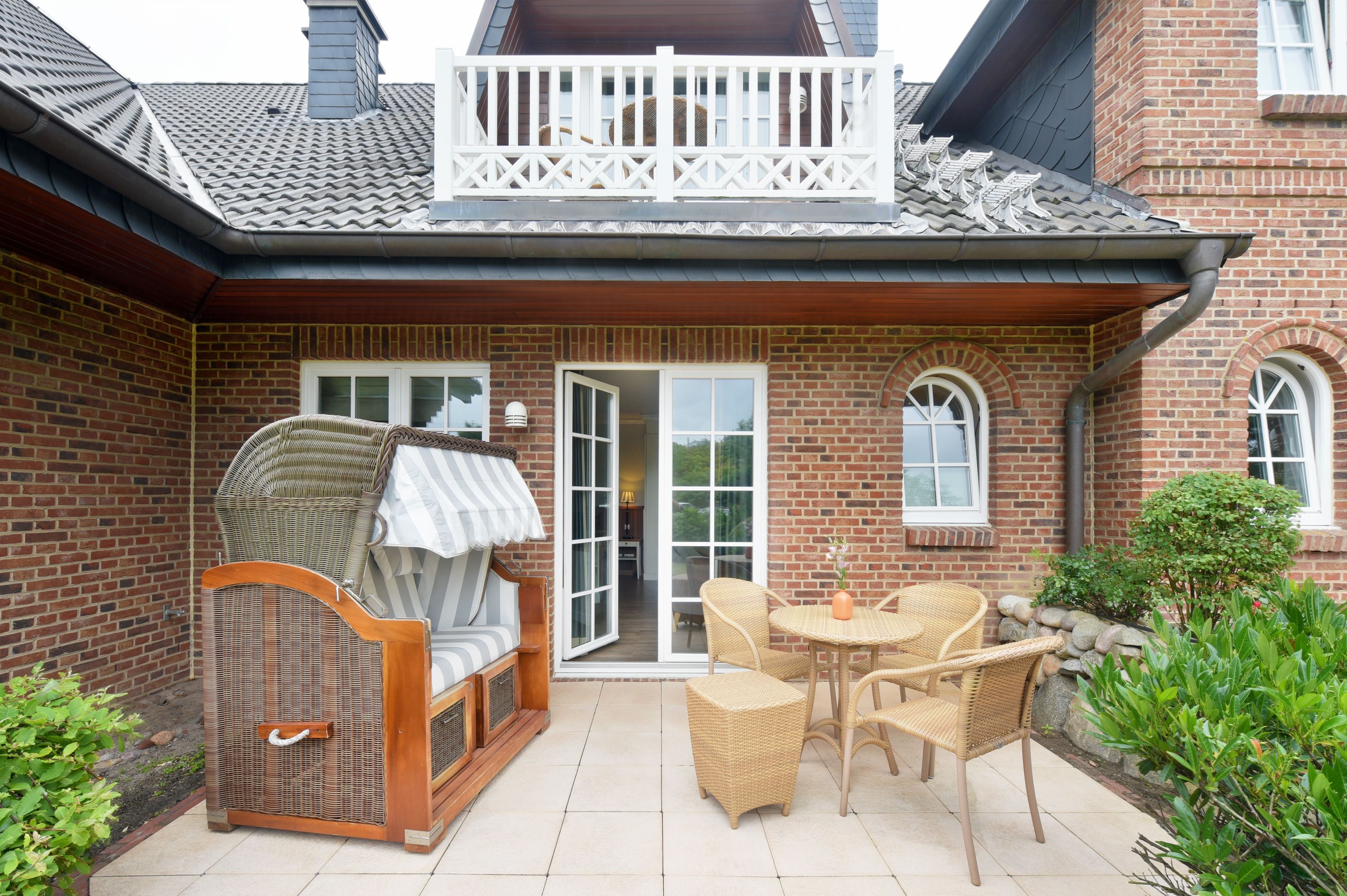 Une maison en briques avec un balcon, un espace extérieur aménagé et couvert avec des meubles en osier, et un petit jardin.
