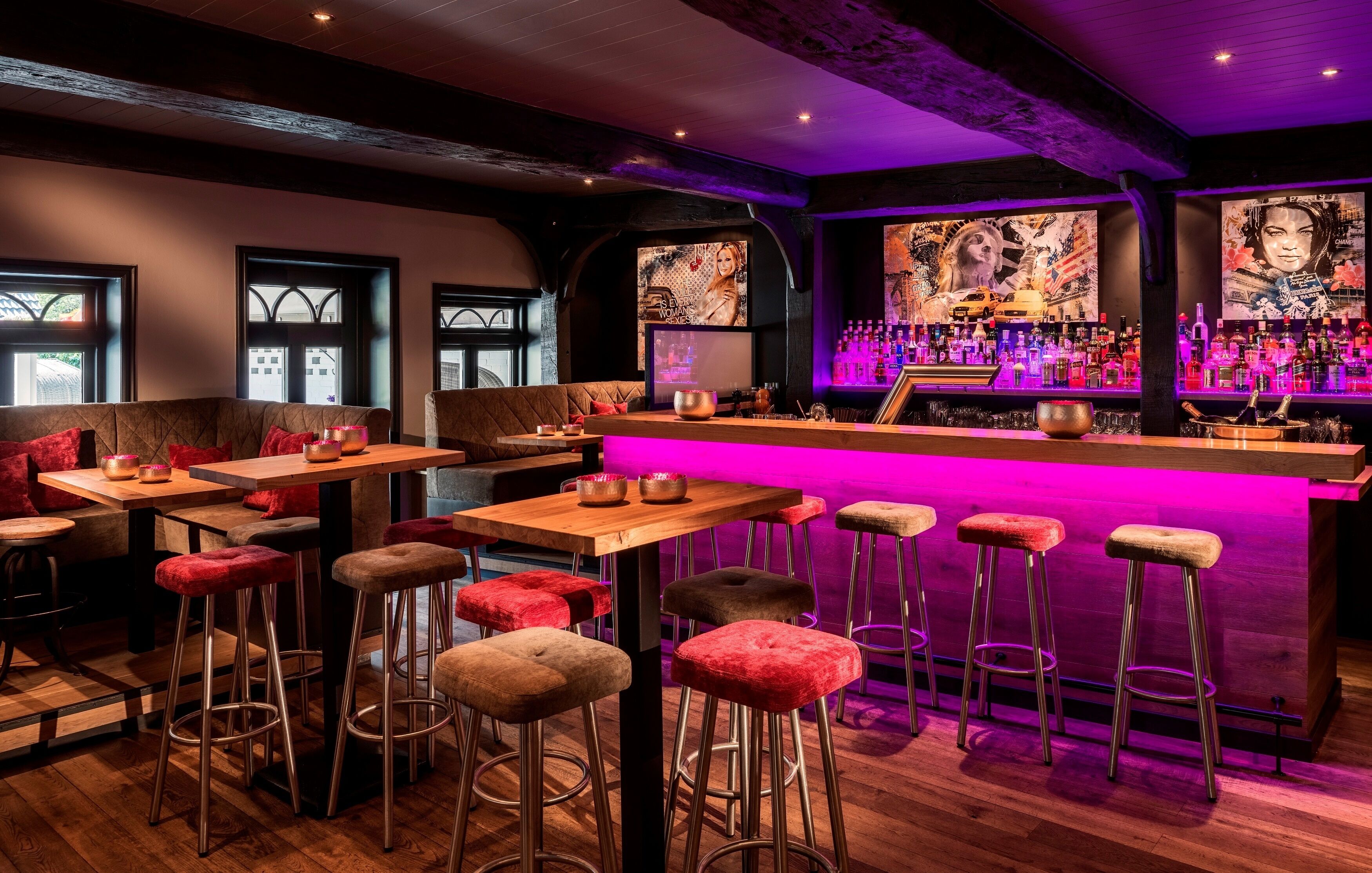 Un espace bar avec des tables en bois et des tabourets rouges rembourrés, un comptoir de bar avec des bouteilles et des œuvres d’art accrochées aux murs.