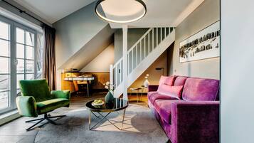 Una sala de estar moderna con un sillón verde, un sofá morado, una mesa de centro redonda y una escalera que sube a un nivel superior.