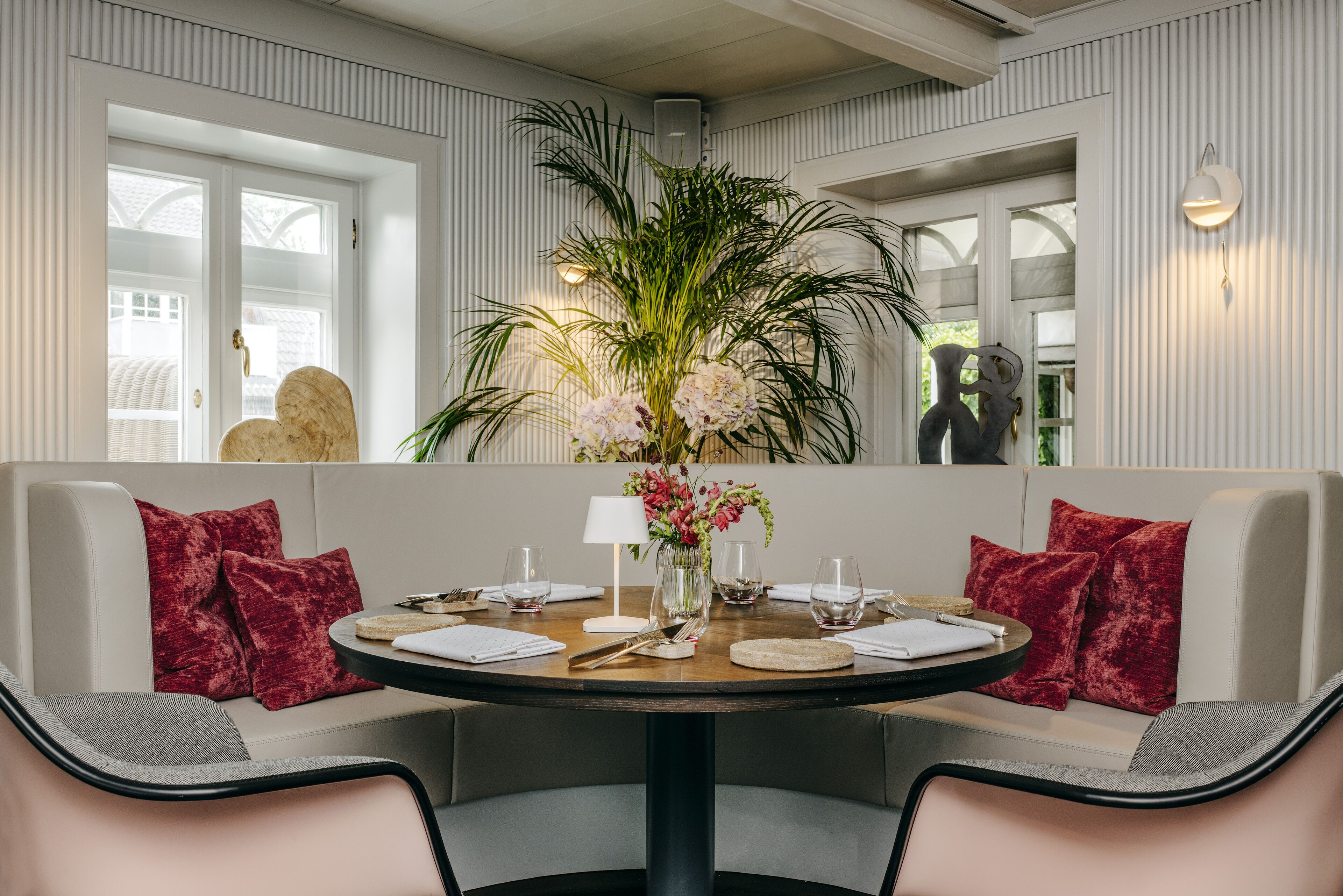 A dining area with a round table set for four, surrounded by two sofas with red cushions, a vase with flowers, and a wall-mounted light fixture.