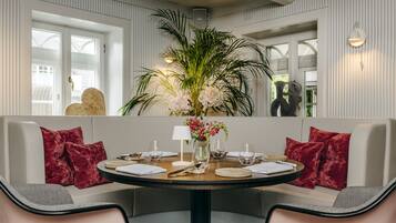 A dining area with a round table set for four, surrounded by two sofas with red cushions, a vase with flowers, and a wall-mounted light fixture.
