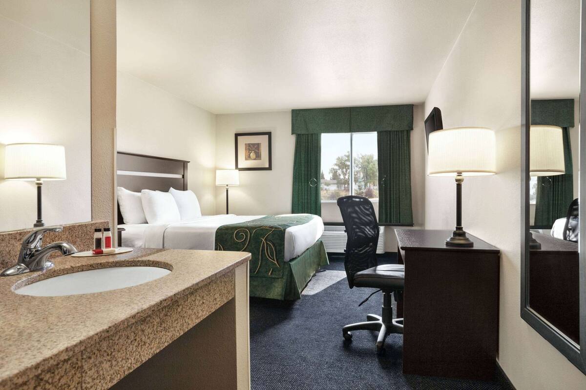A hotel room with a bed, a desk, a chair, a desk lamp, a sink, and a mirror.