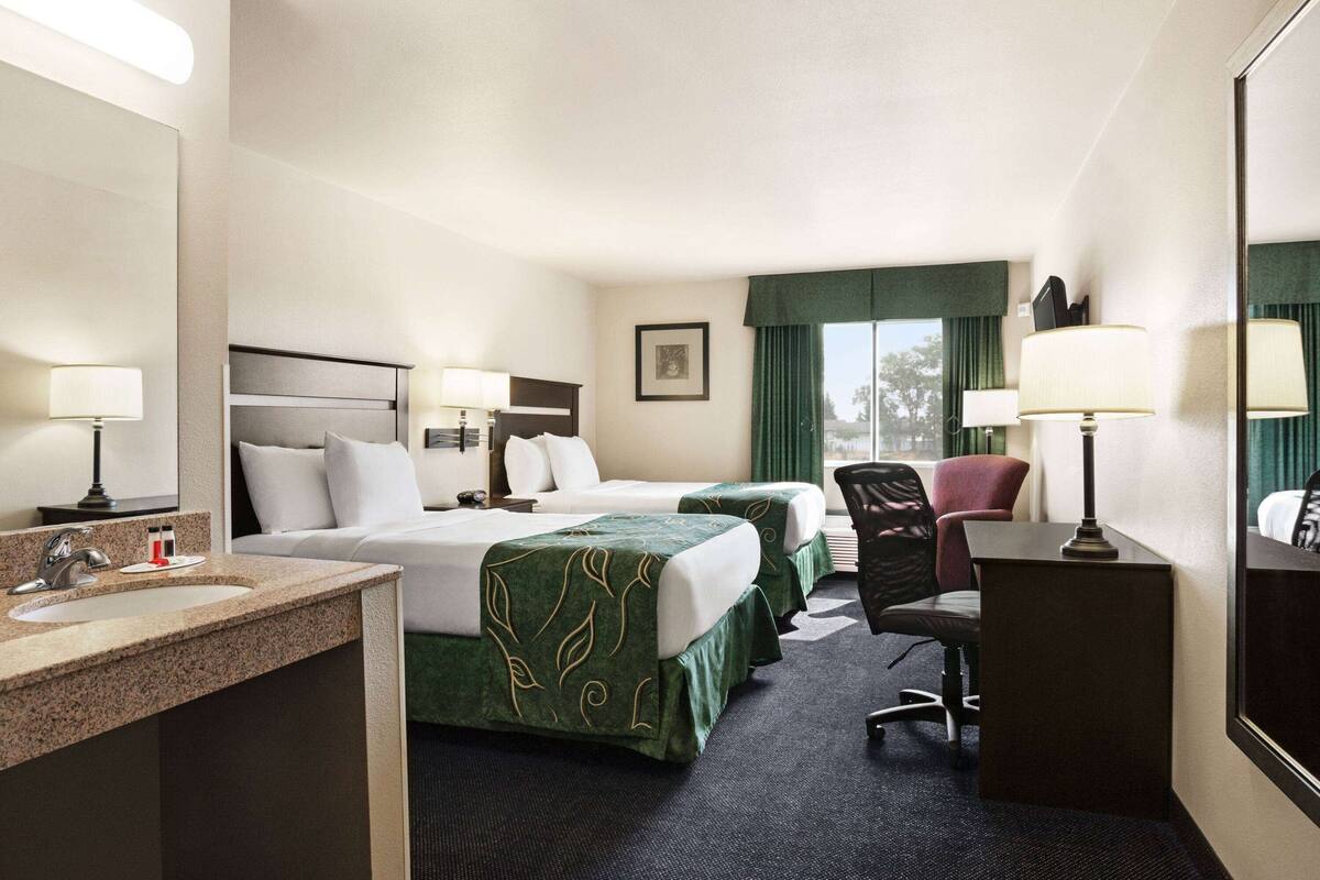 A hotel room with two beds, a desk, a chair, a desk lamp, a mirror, and a sink.