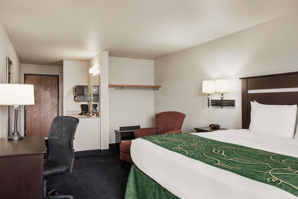 A hotel room with a bed, desk, chair, and a small kitchenette area.