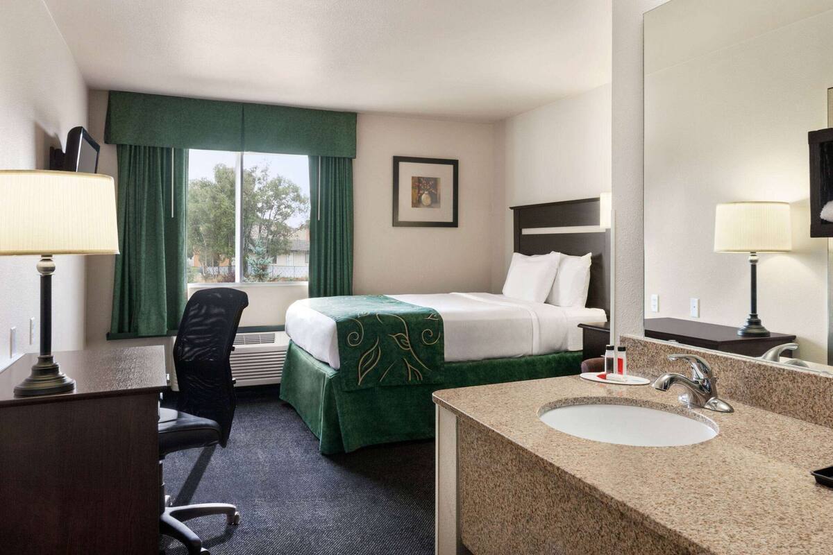 A hotel room with a bed, a desk, a chair, a bathroom with a sink and mirror, and a window with curtains.