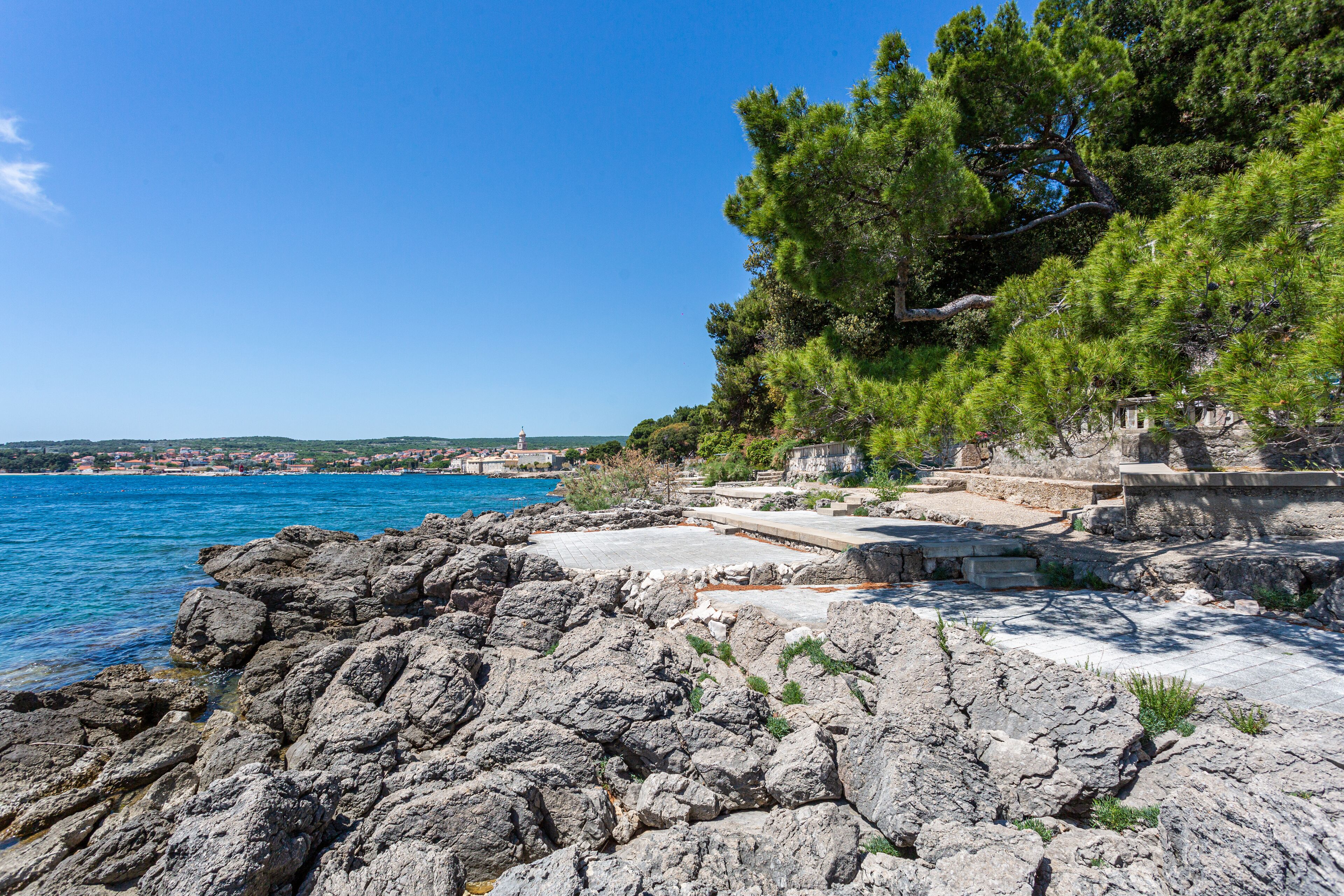 Un'area costiera rocciosa con un mare blu limpido, un sentiero in pietra e vista su una cittadina in lontananza.