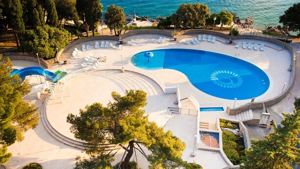 A large swimming pool with a circular design, surrounded by lounge chairs and a landscaped area with trees. - Hotel Drazica (Krk)