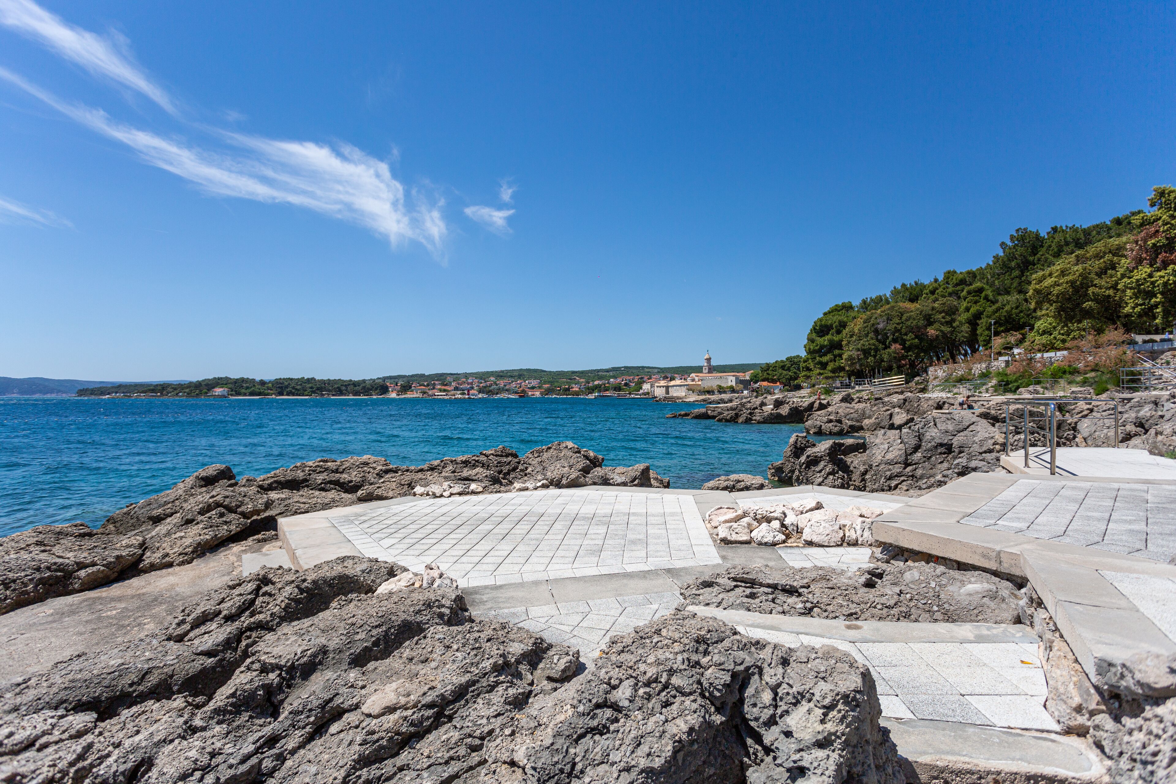 A coastal area with rocky terrain, a paved walkway, and a view of the sea and distant buildings.