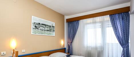 A hotel room with a bed, a desk, a chair, a nightstand, and a wall-mounted lamp.