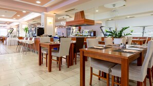 A modern dining area with wooden tables and chairs, a kitchen in the background, and a ceiling with recessed lighting. - Hotel Drazica (Krk)