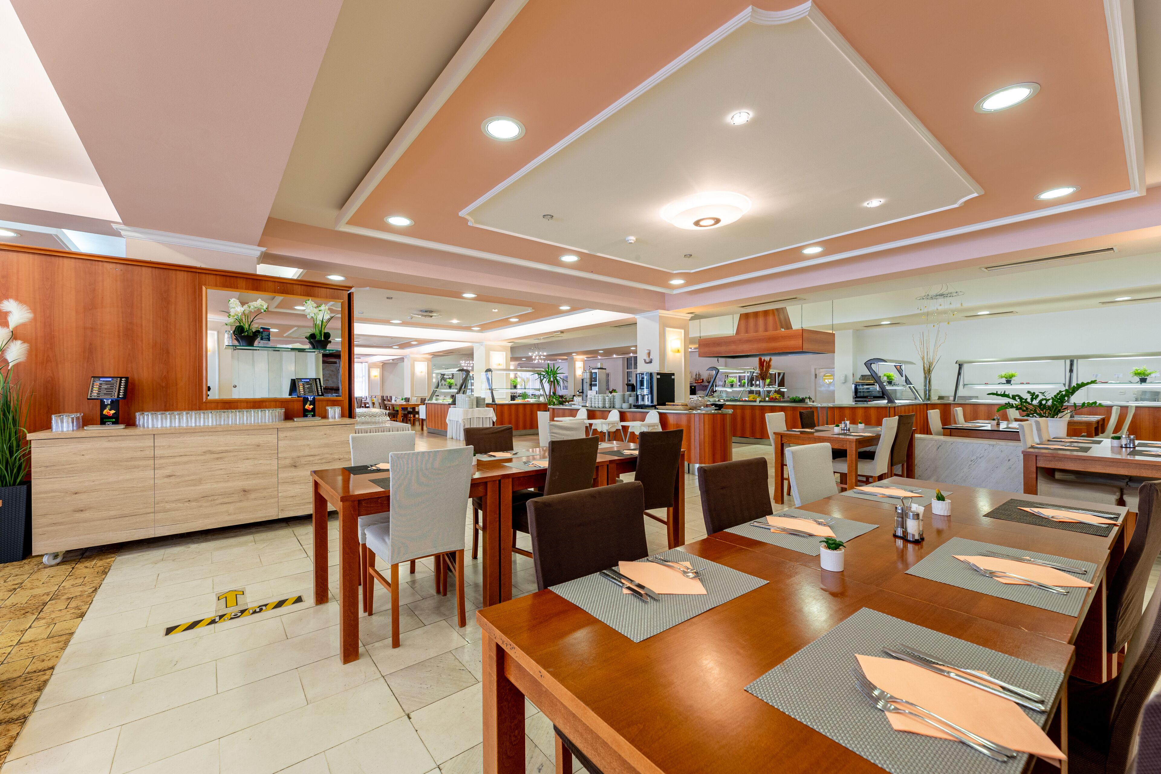 Ein geräumiger Hotel-Speisesaal mit mehreren Tischen und Stühlen, einem Empfangsbereich und einer Küche im Hintergrund.