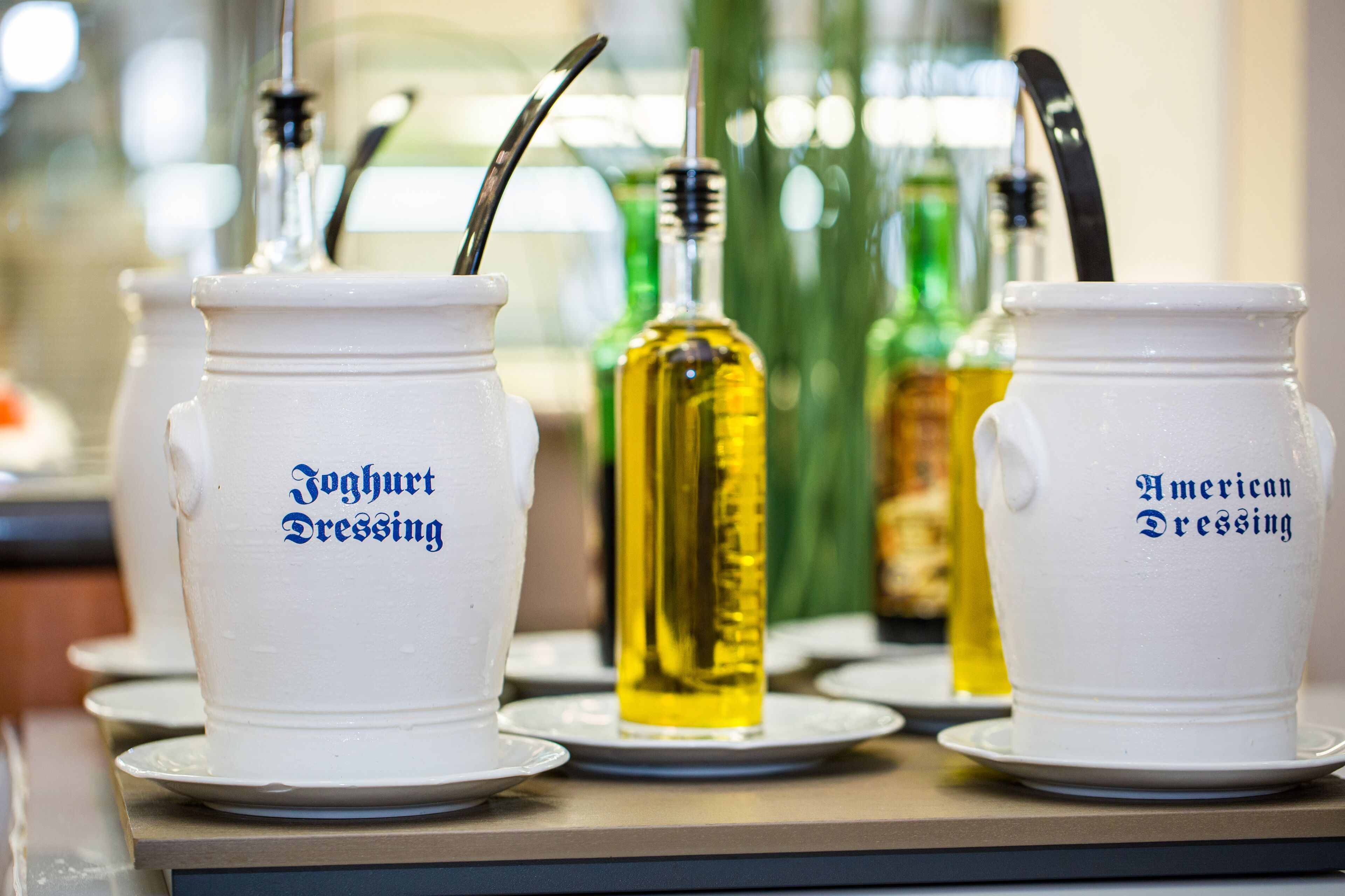 Drei weiße Keramikgießer mit den Aufschriften 'Joghurt Dressing' und 'American Dressing', jeweils mit Strohhalm, auf Untertellern platziert.
