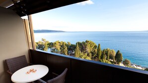A balcony with wicker chairs and a small table, overlooking a body of water and a tree-lined shore. - Hotel Drazica (Krk)